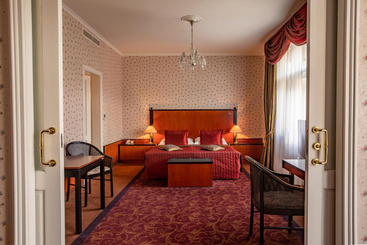 Grandhotel Pupp Karlovy Vary Camera foto The photo shows the interior of a hotel room. Upon entering, you can see a spacious area with a plush, red carpet. The room features a large bed with decorative pillows and a wooden headboard, flanked by bedside tables with lamps. In front of the bed