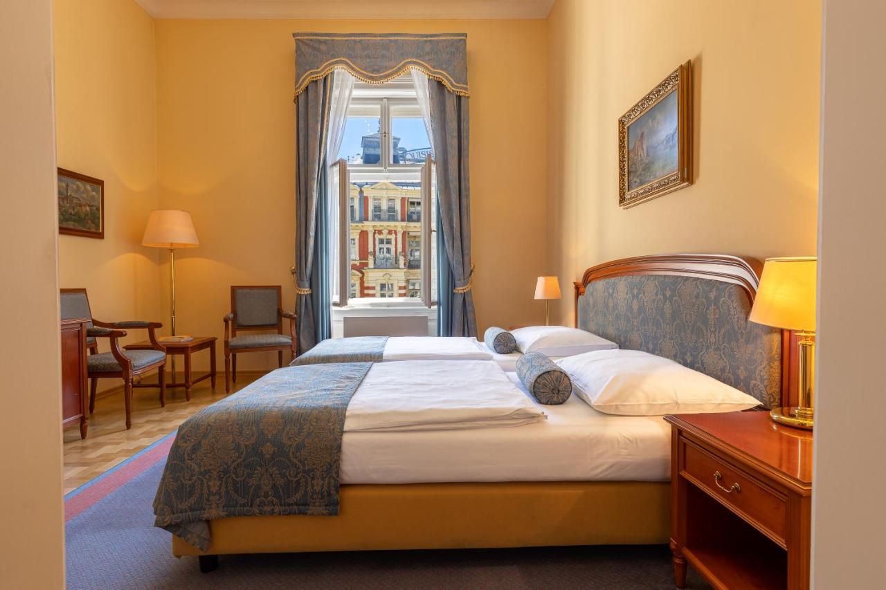 Grandhotel Pupp Karlovy Vary Esterno foto The photo depicts a cozy hotel room or bedroom. It features a neatly made bed with a blue and white color scheme, including decorative pillows. On one side of the bed is a small bedside table with a lamp. The room has warm yellow walls and a large wi