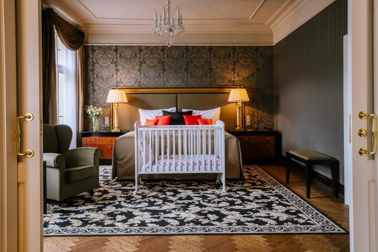 Grandhotel Pupp Karlovy Vary Esterno foto The photo shows a stylish bedroom. The room features an elegant design with a dark patterned wallpaper and a large bed adorned with decorative pillows in red and black. At the foot of the bed is a white crib. There are two bedside lamps providing war
