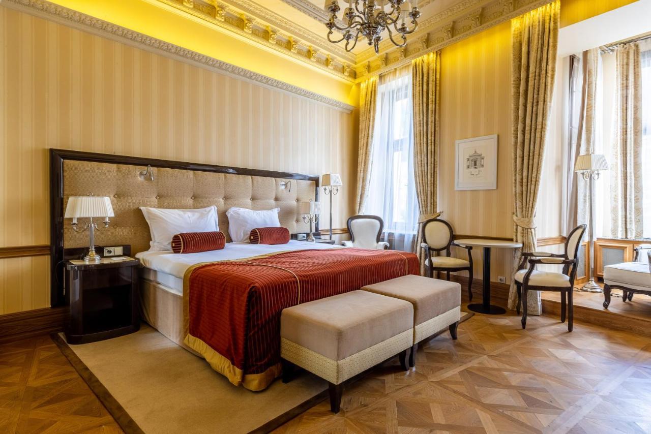Grandhotel Pupp Karlovy Vary Esterno foto The photo shows a luxurious hotel room designed with an elegant interior. The room features a large bed with a tufted headboard, adorned with white bedding and a deep red throw. On either side of the bed, there are bedside lamps and a small seating a