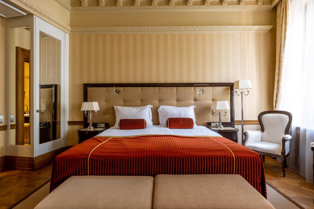 Grandhotel Pupp Karlovy Vary Esterno foto The photo shows an elegantly decorated hotel room featuring a large bed with a plush headboard. The bed is dressed in rich, orange bedding, complemented by white pillows and decorative cushions. Flanking the bed are two bedside lamps on matching nigh