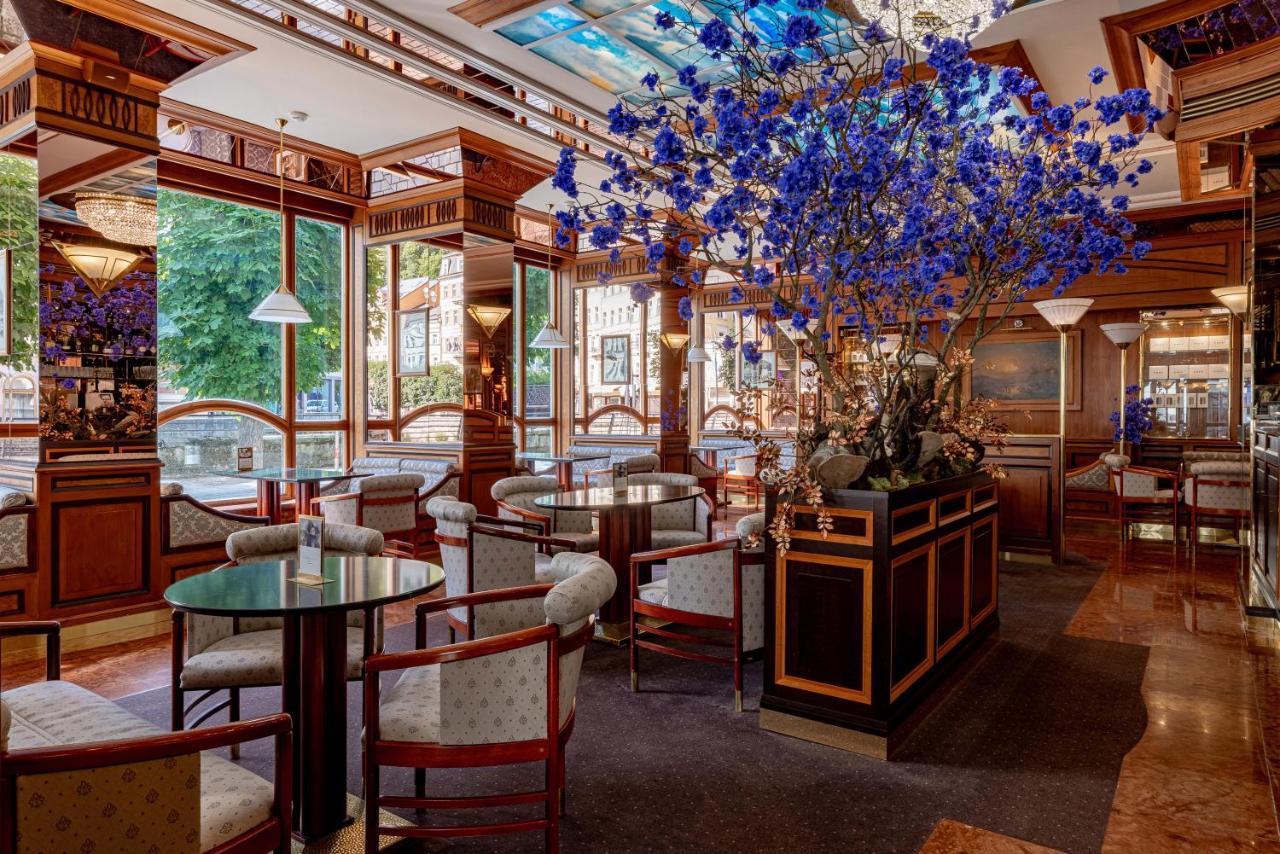 Grandhotel Pupp Karlovy Vary Esterno foto The photo shows an elegant interior of a restaurant or café. It features wooden furnishings with intricate details, including circular tables and upholstered chairs. A large floral arrangement, predominantly featuring blue flowers, is placed in the c