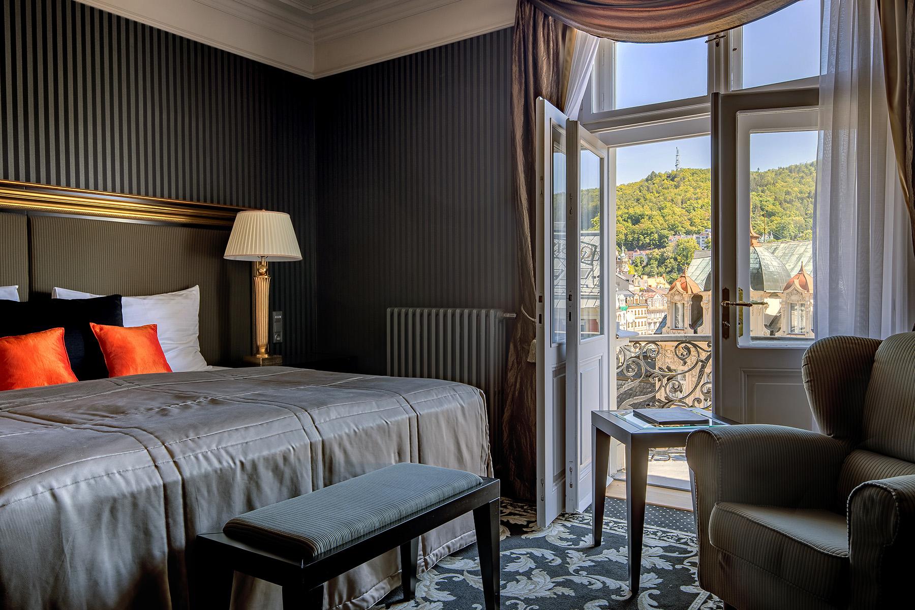 Grandhotel Pupp Karlovy Vary Esterno foto The photo shows a cozy hotel room with a sophisticated design. The room features a bed with a plush grey duvet and decorative pillows in black and orange. There is a bedside lamp beside the bed. A comfortable armchair sits in one corner. The room has