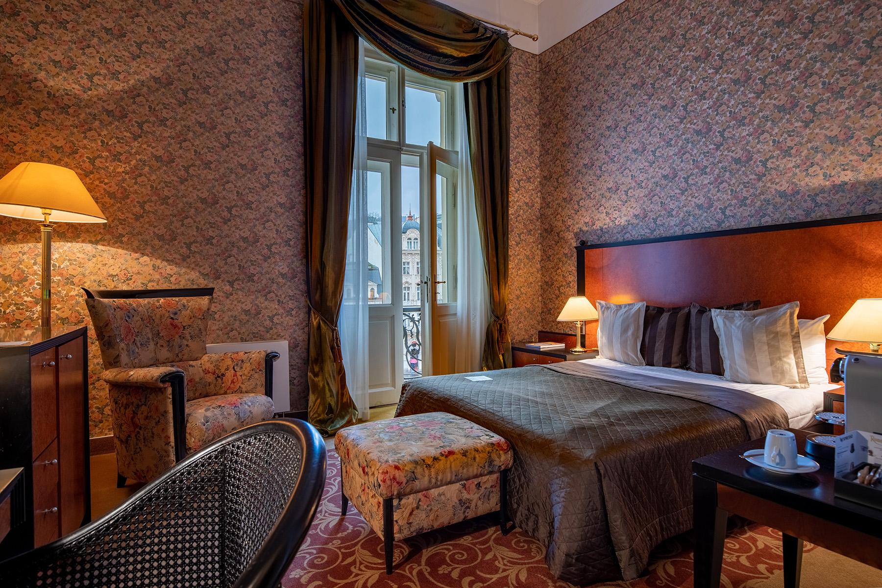 Grandhotel Pupp Karlovy Vary Esterno foto The photo shows a cozy hotel room featuring a queen-sized bed with dark bedding and several decorative pillows. There is a comfortable armchair and an ottoman in a floral pattern, adding a touch of elegance. A wooden bedside table holds a lamp and a 