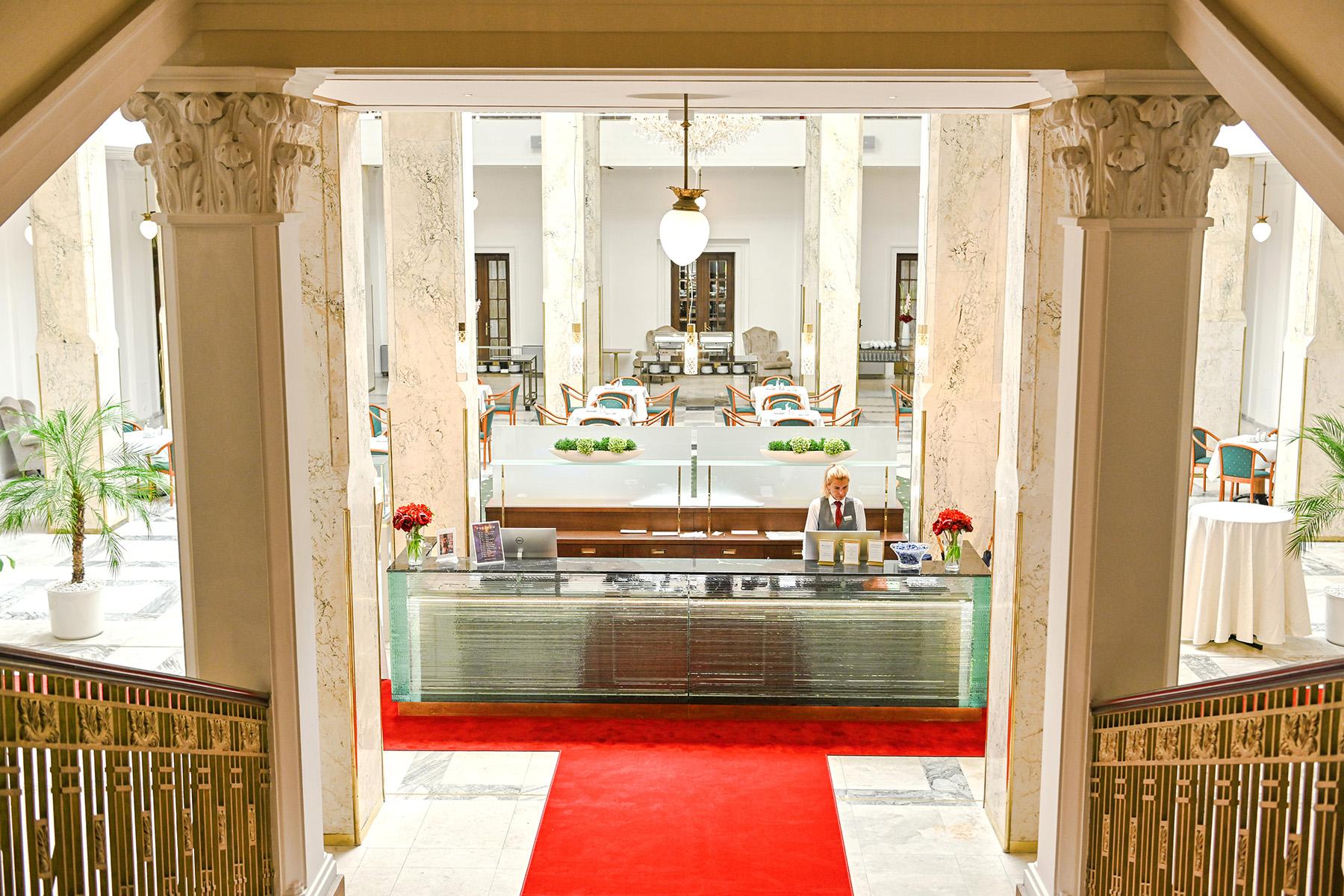 Grandhotel Pupp Karlovy Vary Esterno foto The photo shows the interior of a hotel lobby. There is a reception desk made of glass and wood, situated at the front, with a staff member standing behind it. The lobby features elegant columns and a high ceiling, giving it a grand appearance. A red