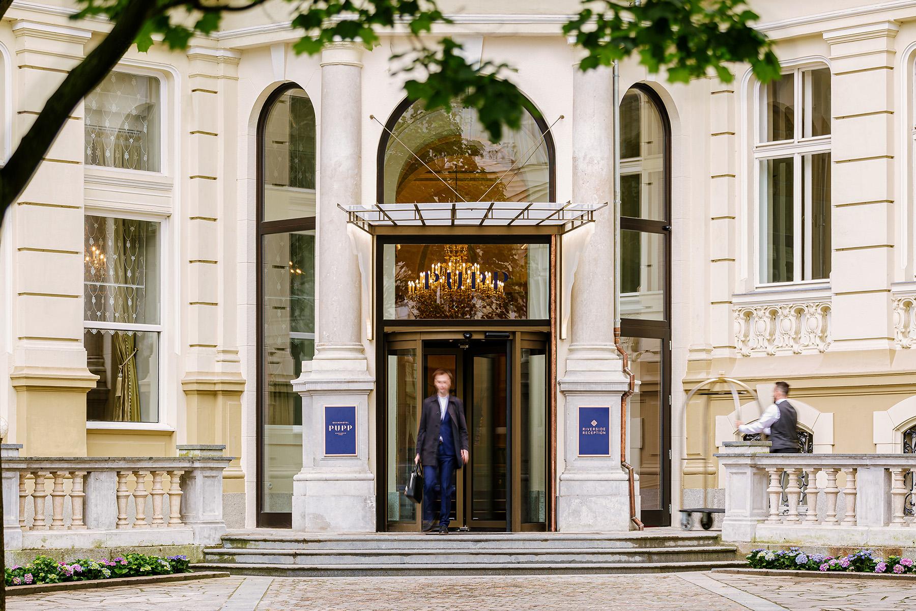 Grandhotel Pupp Karlovy Vary Esterno foto The photo shows an elegant entrance to a building, likely a hotel or a high-end venue. There are large, grand pillars supporting an awning over the entrance. A chandelier is visible inside, suggesting a luxurious interior. A man in formal attire is w