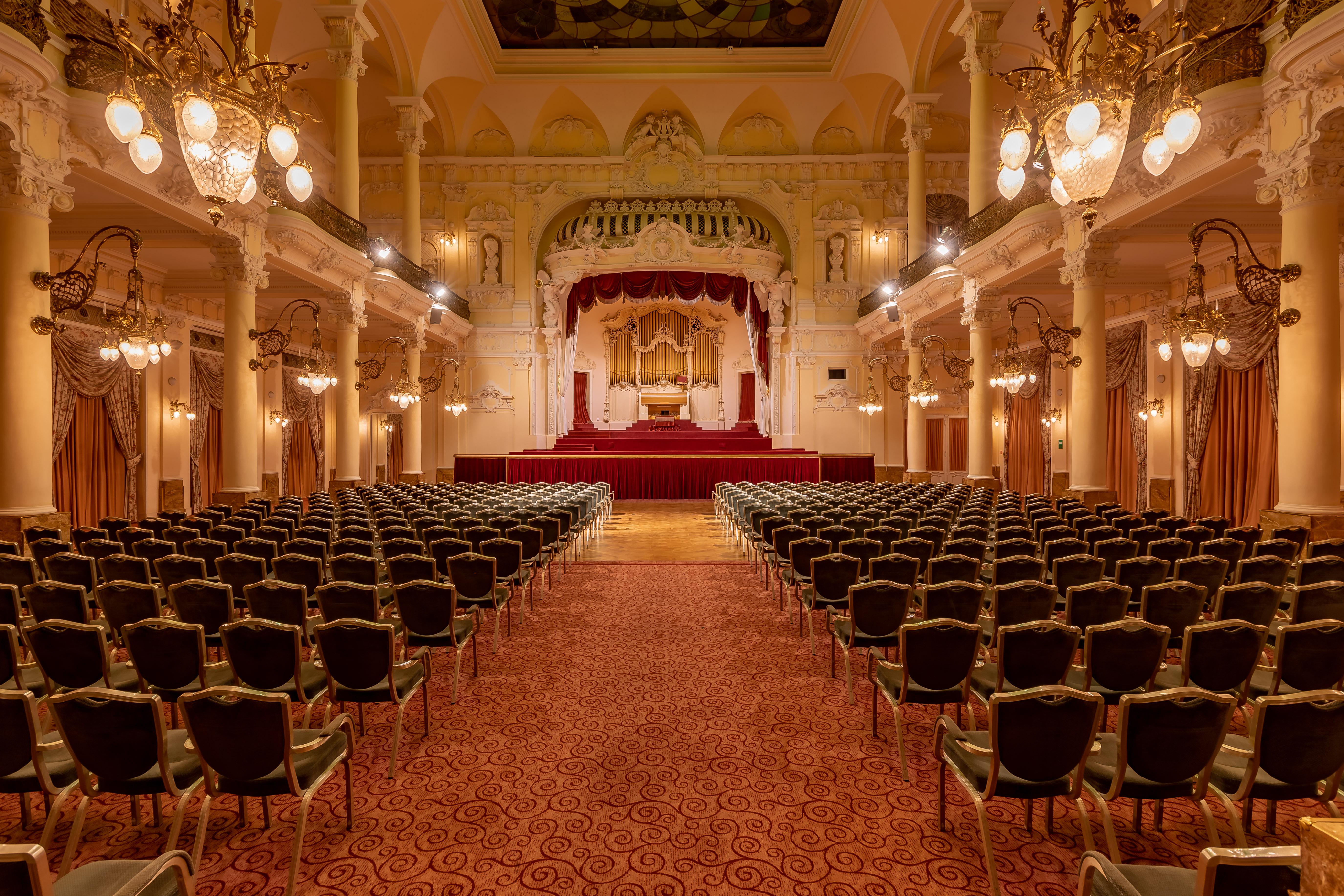 Grandhotel Pupp Karlovy Vary Esterno foto The photo showcases an elegant interior of a theater or concert hall. It features a grand stage in the background, adorned with red curtains and decorative elements. The seating area is filled with rows of green upholstered chairs arranged in a symme