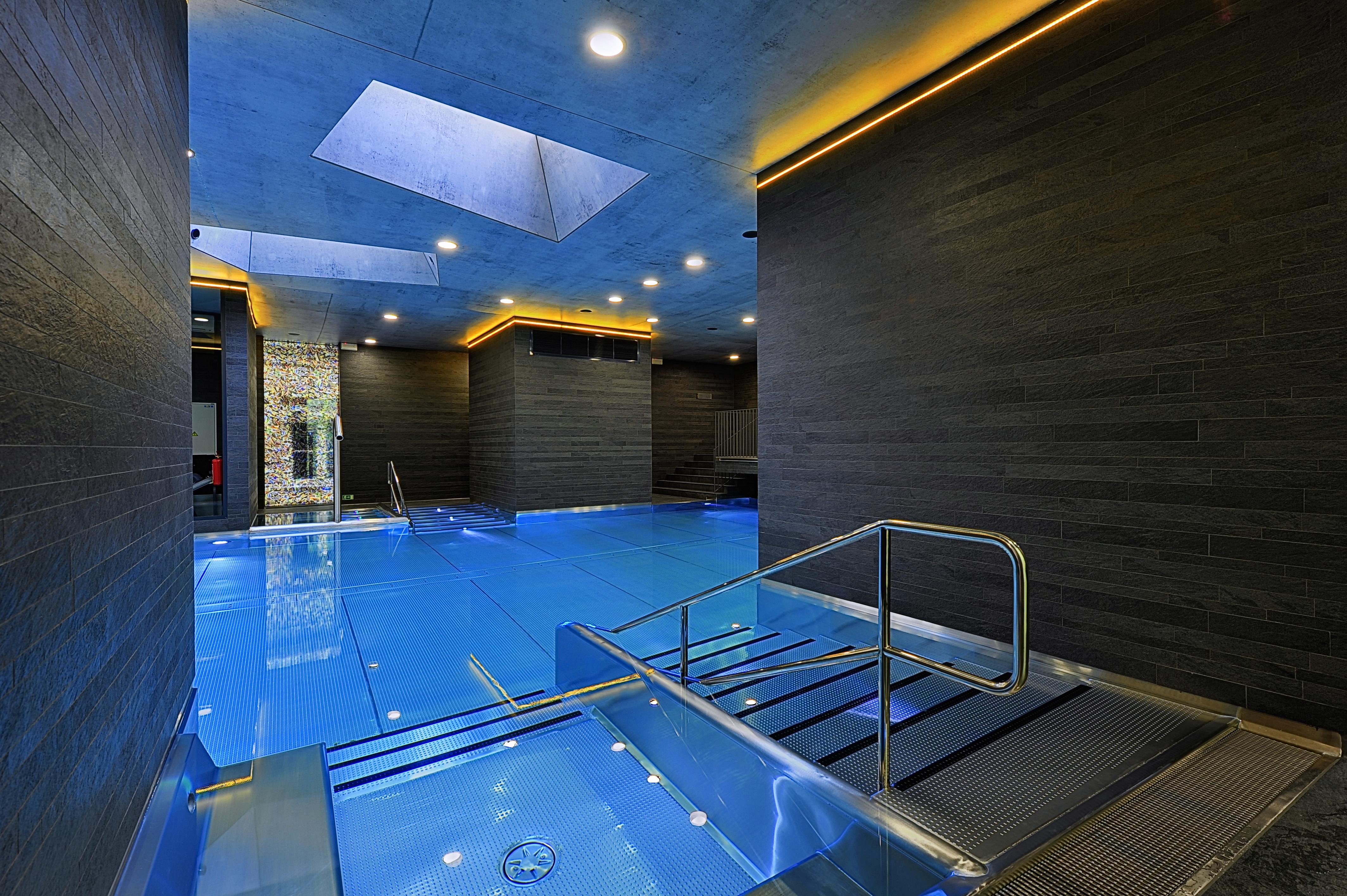 Grandhotel Pupp Karlovy Vary Esterno foto The photo features a modern indoor swimming pool area. The pool is designed with a sleek, contemporary aesthetic, featuring a vibrant blue water surface. The surrounding walls are made of dark materials, possibly stone or wood, complemented by warm l