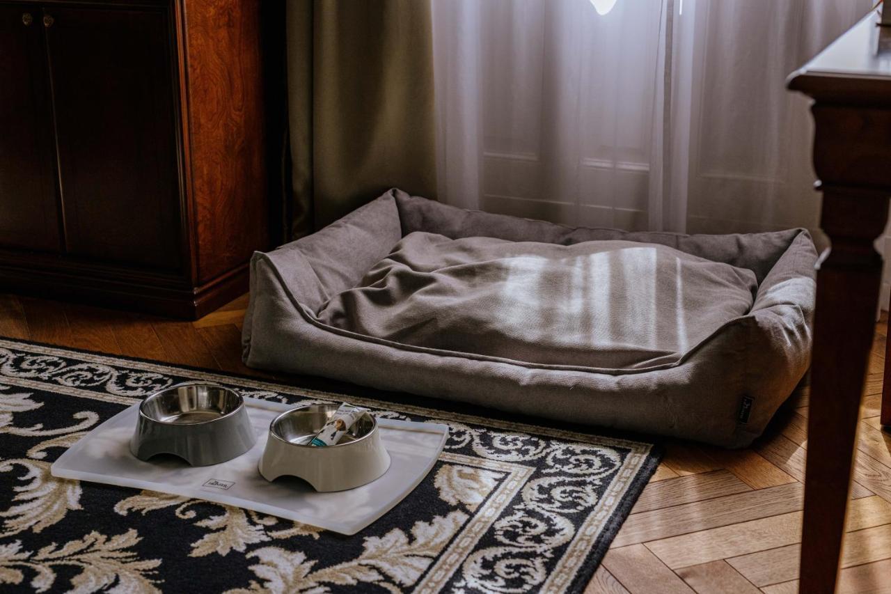 Grandhotel Pupp Karlovy Vary Esterno foto The photo shows a cozy pet area inside a room. There is a soft, rectangular pet bed in a muted color, positioned against a wall, with some light filtering through a window. In front of the bed, there are two pet bowls placed on a mat, one bowl is gre