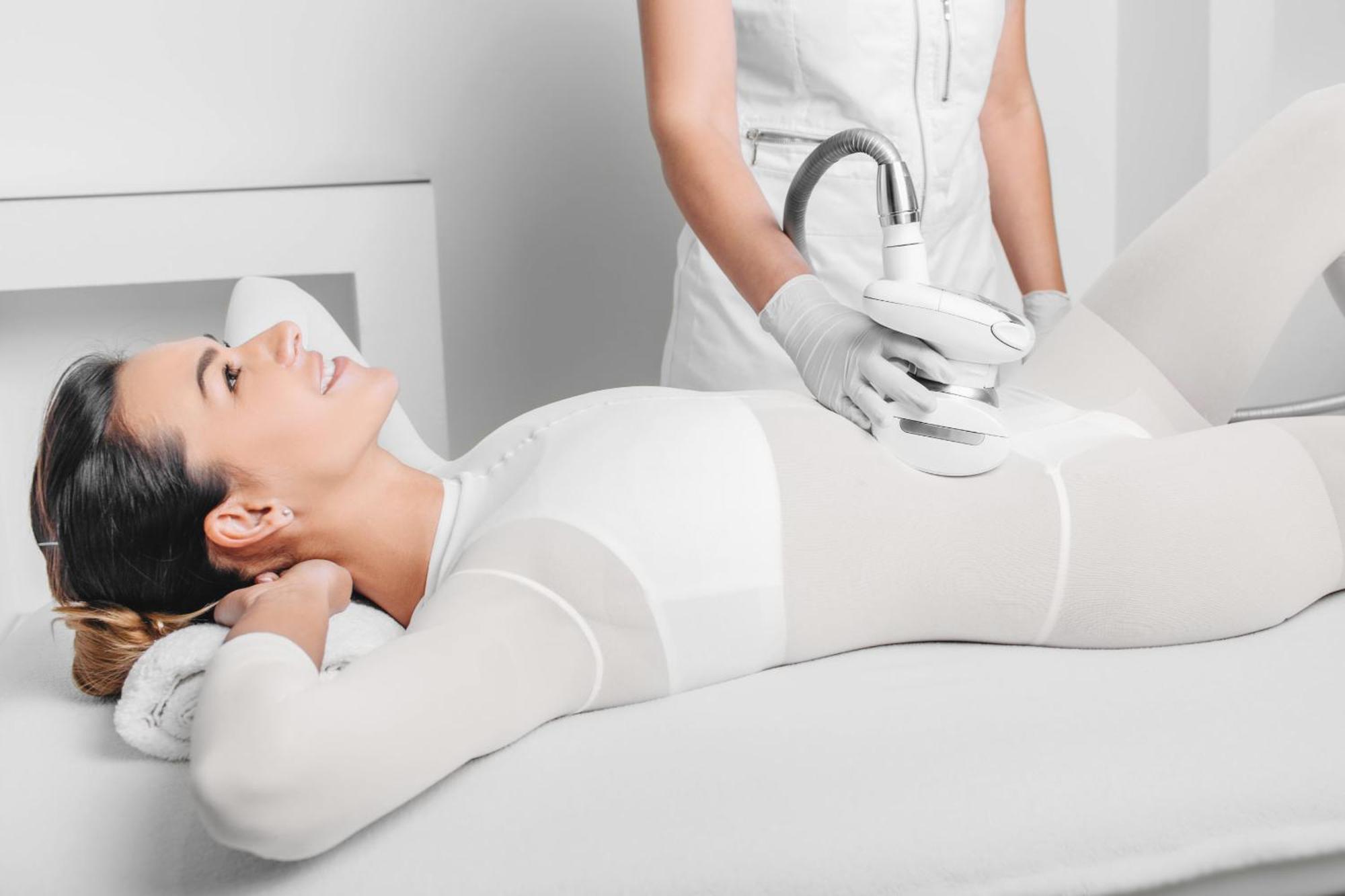 Grandhotel Pupp Karlovy Vary Esterno foto The photo depicts a woman lying on a treatment table, appearing relaxed and smiling. She is wearing a form-fitting white outfit, likely designed for a cosmetic or wellness procedure. A technician, dressed in a white outfit and gloves, is applying a d