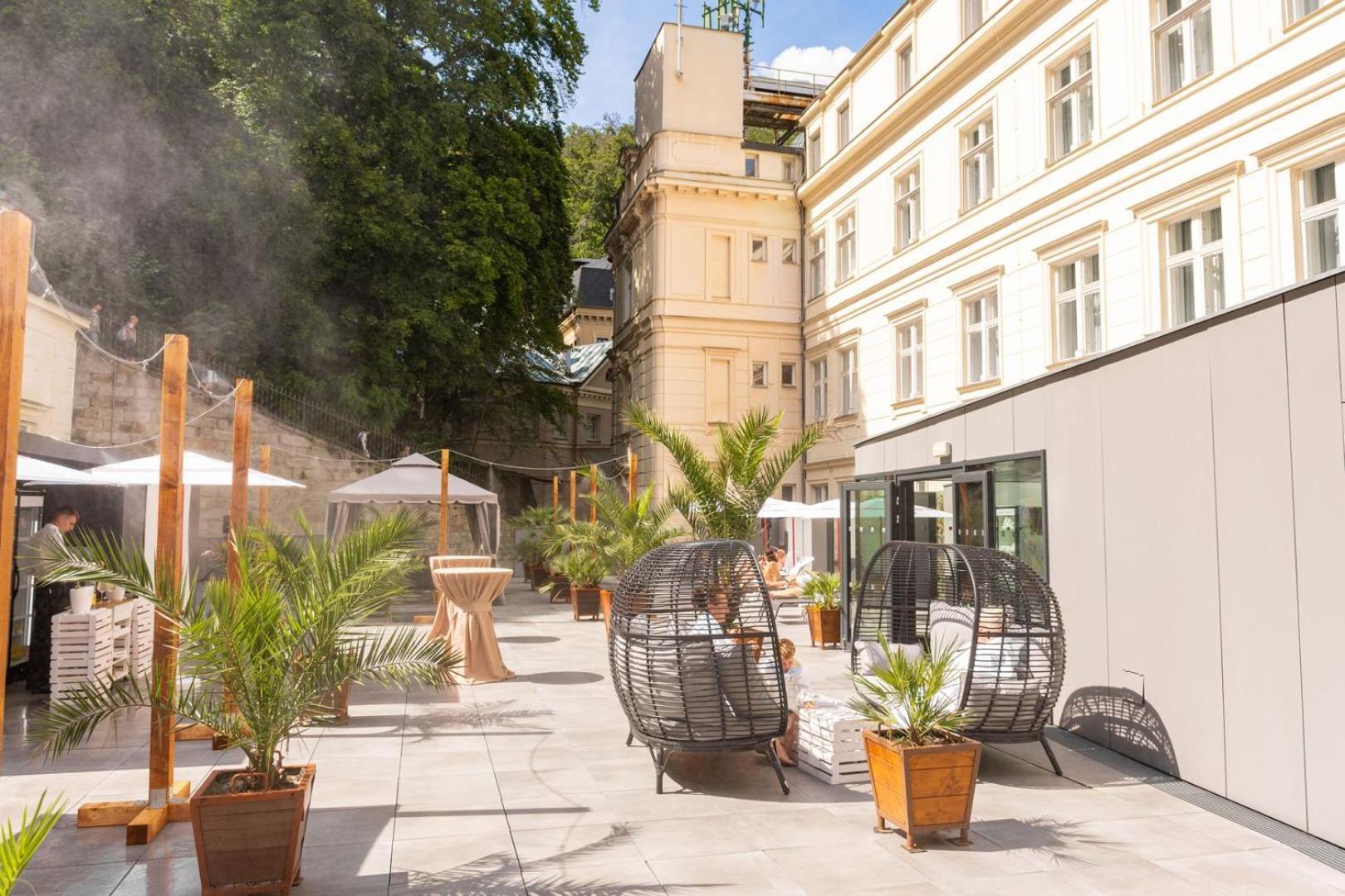 Grandhotel Pupp Karlovy Vary Esterno foto The photo shows a stylish outdoor area with a mix of modern and classic architecture. There are several large potted plants, likely palm trees, which add a tropical feel to the space. Two round, open wicker chairs are placed on a paved surface, provi